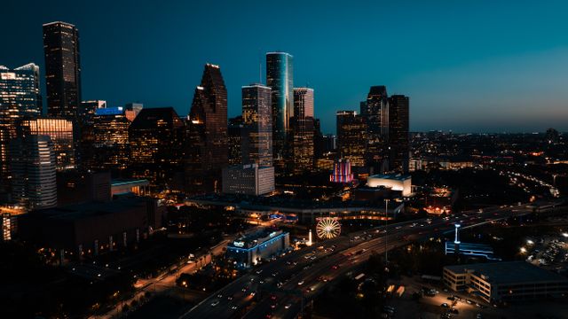 A city skyline at dusk features tall, illuminated buildings, busy highways with moving cars, and a lit-up Ferris wheel in the foreground under a darkening blue sky.