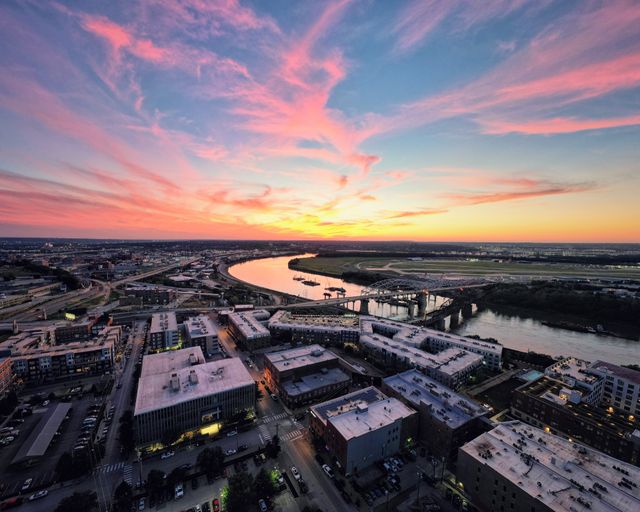 Aerial view of a city at sunset with pink clouds, buildings in the foreground, and a winding river reflecting the colorful sky.