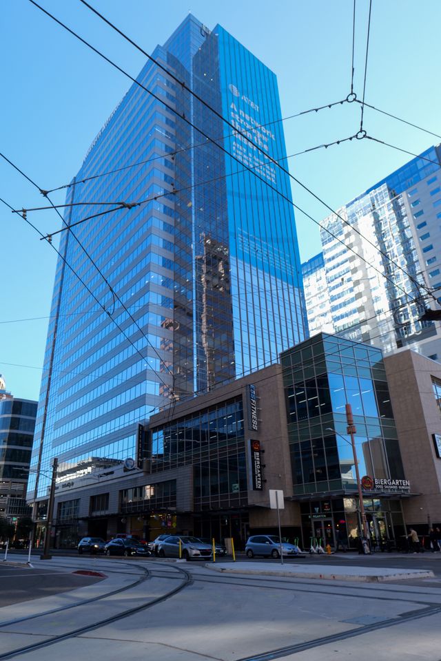 Tall blue glass skyscraper rises behind a shorter modern building with shops and restaurants. Streetcar wires cross above city streets with cars below. Clear blue sky in the background.