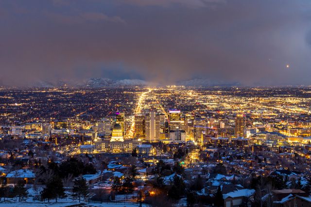Cityscape at dusk showing a brightly lit downtown area with snow-covered buildings in the foreground and mountains under a cloudy sky in the background.