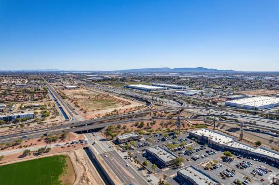 Aerial view of a cityscape with highways, commercial buildings, farmland, and distant mountains under a clear blue sky.