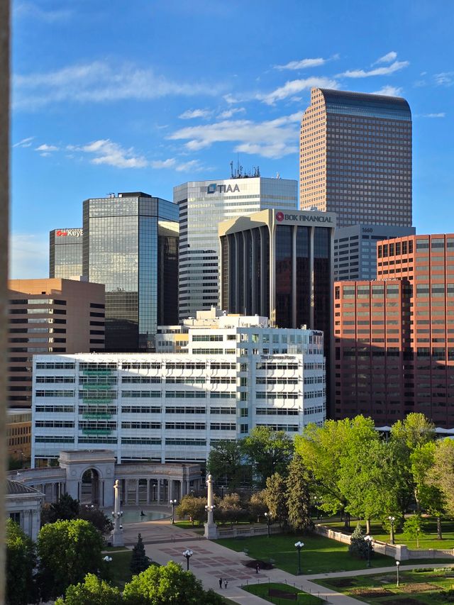 Downtown cityscape with tall modern office buildings, blue sky, and lush green park with trees in the foreground.