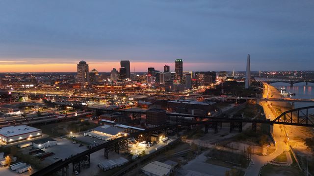 A city skyline at dusk, with buildings lit up and a river on the right. The sky is blue and orange, and the Gateway Arch is visible near the riverbank, with bridges crossing the water.